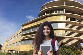 Estudiante sonriente frente al edificio emblemático del instituto, mostrando la entrada principal en un día soleado
