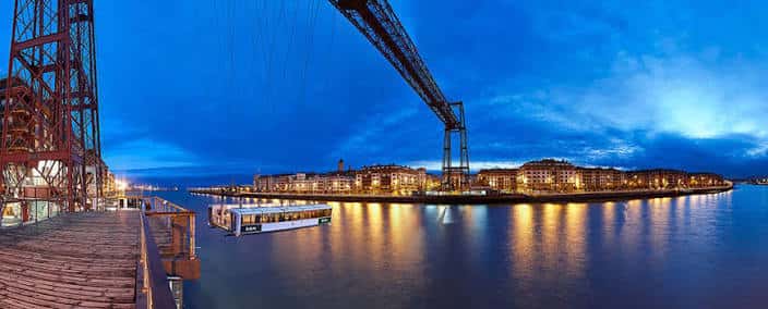 Puente Bizkaia iluminado al atardecer con reflejos en el agua y embarcación turística