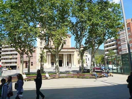 Plaza arbolada en un día soleado con personas caminando y la fachada clásica de la iglesia