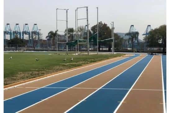 Pista de atletismo con carriles azules y marrones, vista desde la curva con grúas portuarias al fondo