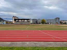 Vista panorámica de las pistas de atletismo rojas con el estadio y edificios urbanos al fondo bajo un cielo nublado
