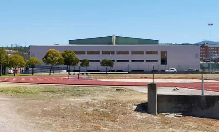 Amplia vista exterior del complejo deportivo con pista roja, césped seco y edificio blanco con techo verde