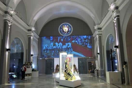 Salón central del museo con vitrinas de bordados y pantalla digital