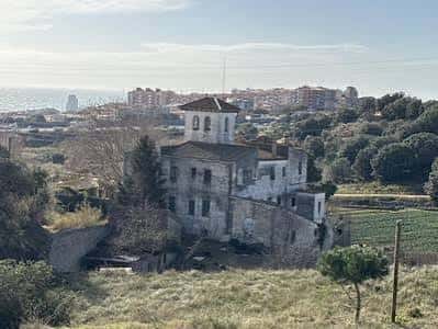 Panorámica de Can Portell Vell desde una colina, mostrando el edificio rodeado de vegetación y la ciudad de Mataró al fondo