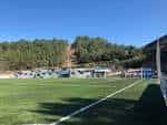 Vista panorámica del Campo de Fútbol Municipal Os Carrís con césped artificial y gradas bajo cielo azul.