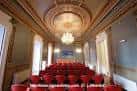 Salón de actos con butacas rojas, escenario y techo ornamentado