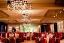 Salón de eventos preparado con mesas y sillas vestidas de rojo, y elegantes lámparas de araña.