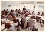Clase con alumnos sentados en pupitres y un profesor al fondo
