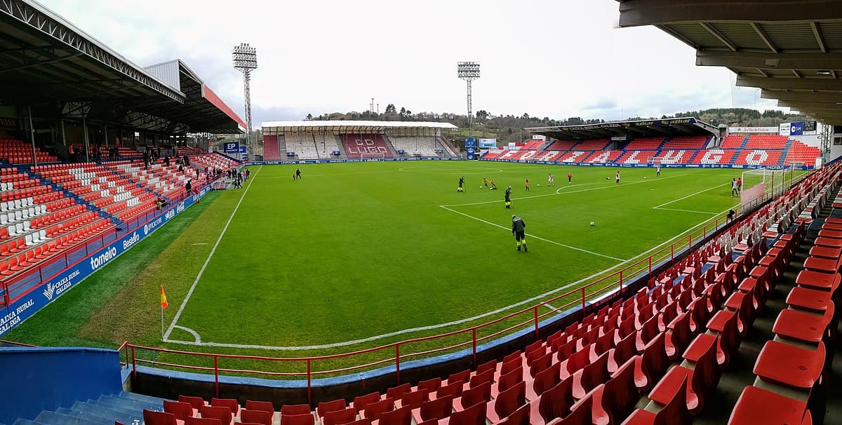 Vista interior del Estadio Anxo Carro con asientos rojos y campo de fútbol.