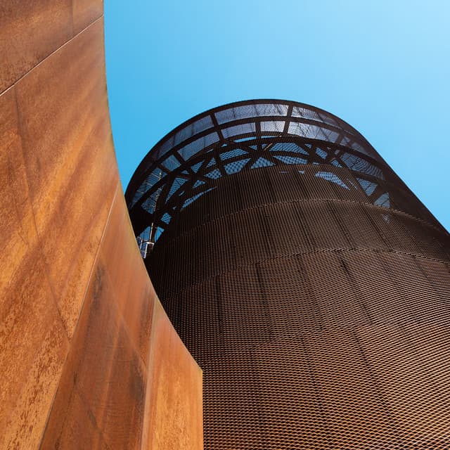 Perspectiva ascendente de una torre cilíndrica de acero corten bajo cielo azul