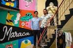 Dos personas posando junto a un mural colorido y una estatua en la escalera de Galería Maraca