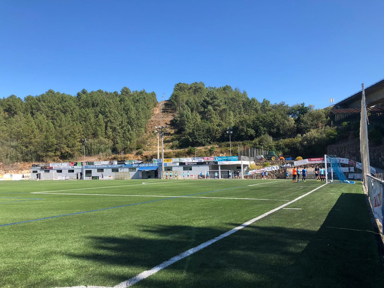 Vista panorámica del Campo de Fútbol Municipal Os Carrís con césped artificial y gradas bajo cielo azul.