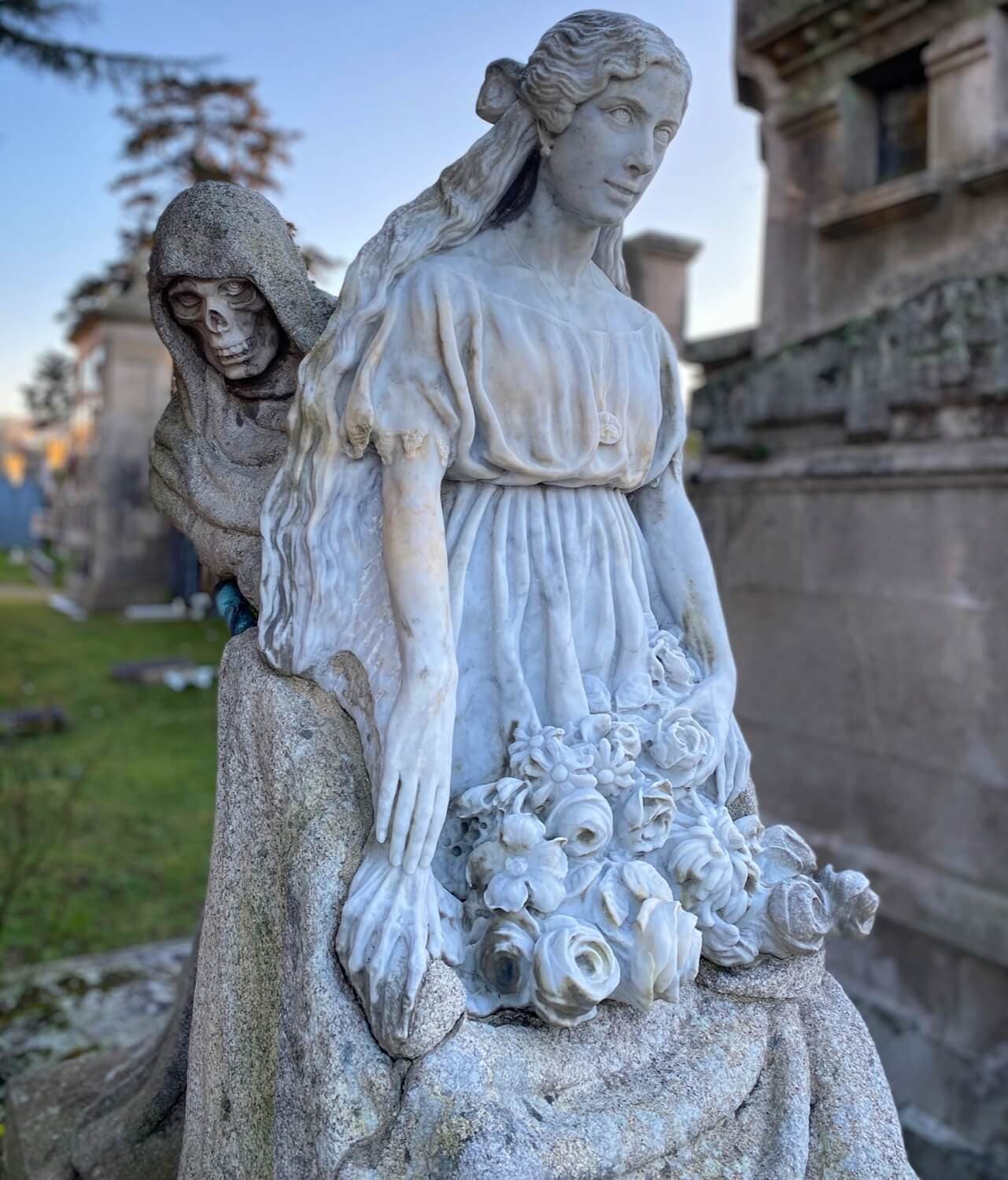 Estatua de mujer con flores y figura encapuchada en el cementerio